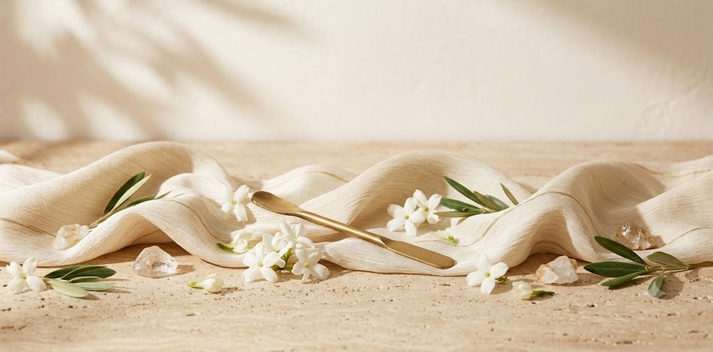A styled flat-lay of a cream-colored linen fabric draped on a sandy surface, accented with white flowers, green leaves, and a gold spoon. 